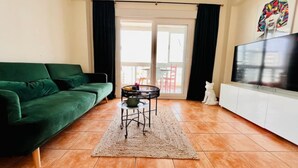 Una sala de estar con un sofá verde, una mesita redonda con un cuenco, un mueble blanco con un televisor y un ventanal corredero que da a un espacio exterior.