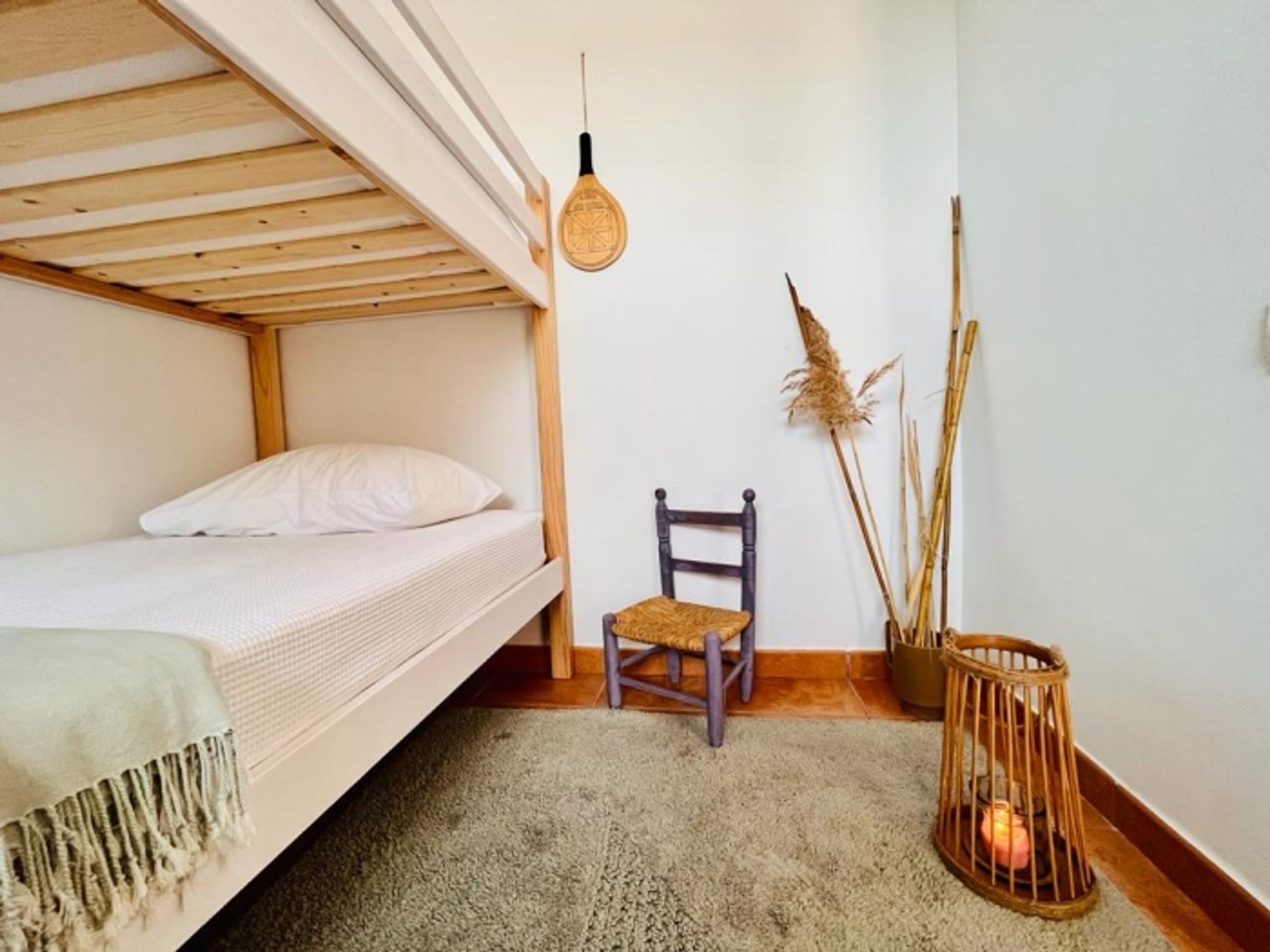 Un lit superposé avec un matelas blanc et un seul oreiller, une chaise en bois avec un siège en osier, un luminaire suspendu et une bougie dans un panier tressé.