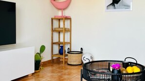Una sala de estar con televisor, estante con una decoración de labios rosados, un cuadro, una cesta negra con frutas y una planta en maceta.