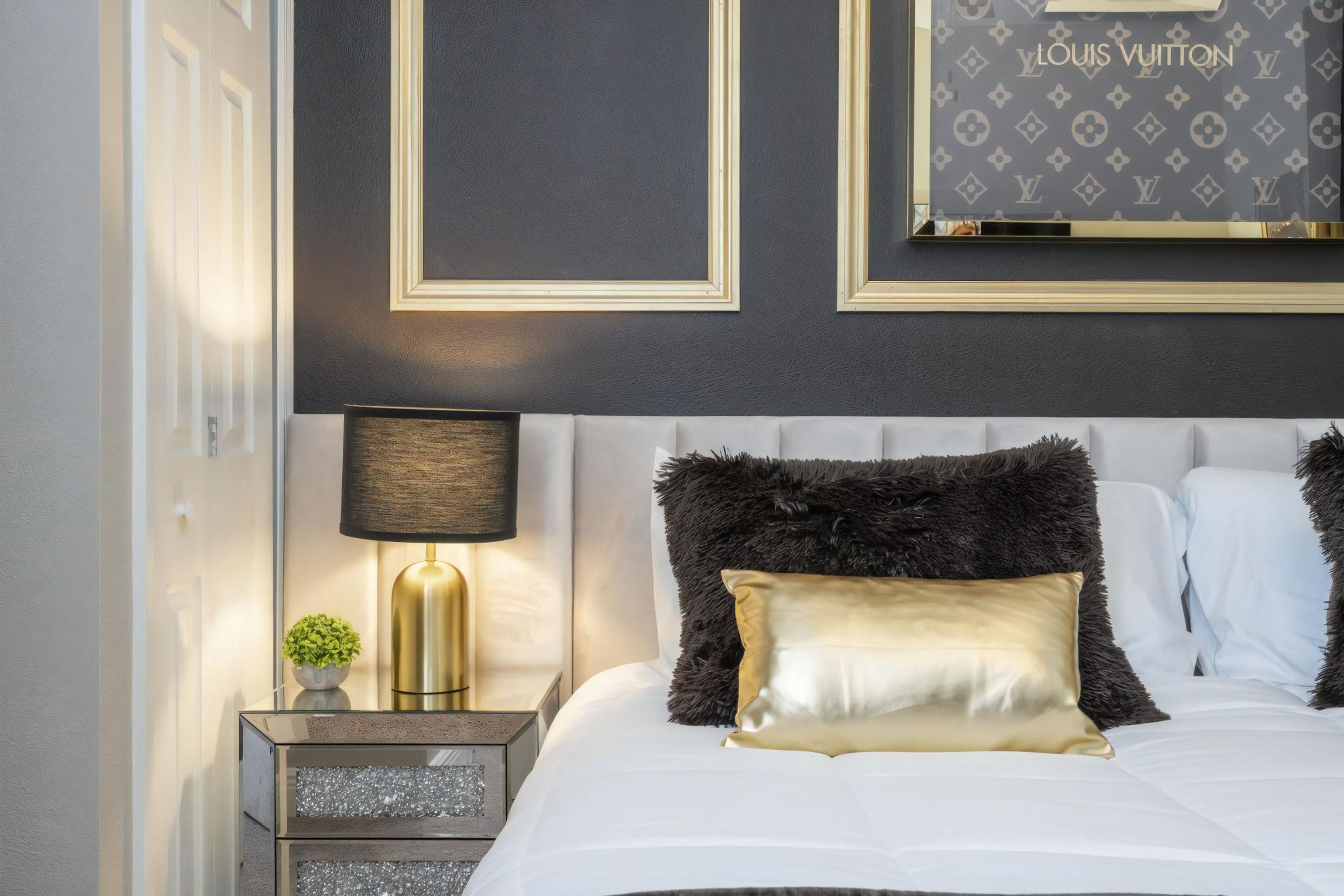 Une chambre moderne avec un lit blanc, des oreillers moelleux de couleur foncée, un coussin décoratif doré, une lampe dorée et une œuvre d’art encadrée de Louis Vuitton accrochée au mur.