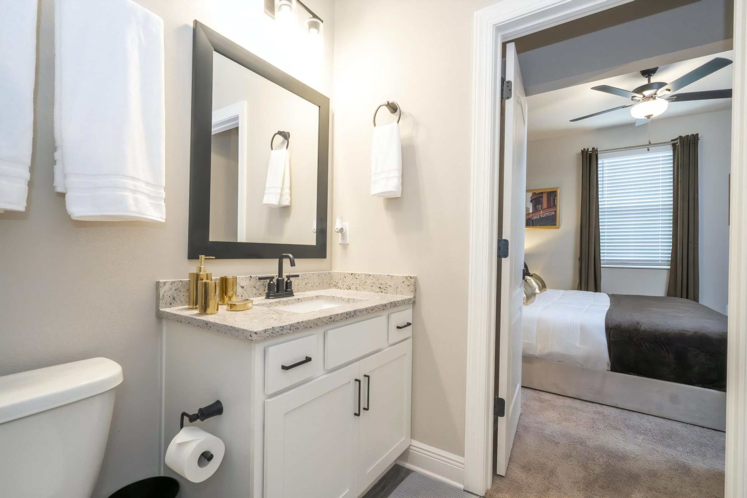 A bathroom with a white toilet, a sink with gold fixtures, and a mirror. There is a bed visible through an open door in the adjacent room.
