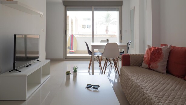 Un salon moderne doté d’un téléviseur à écran plat, d’un canapé avec des coussins rouges, d’une table à manger avec des chaises et d’un balcon où se trouve une planche de surf.