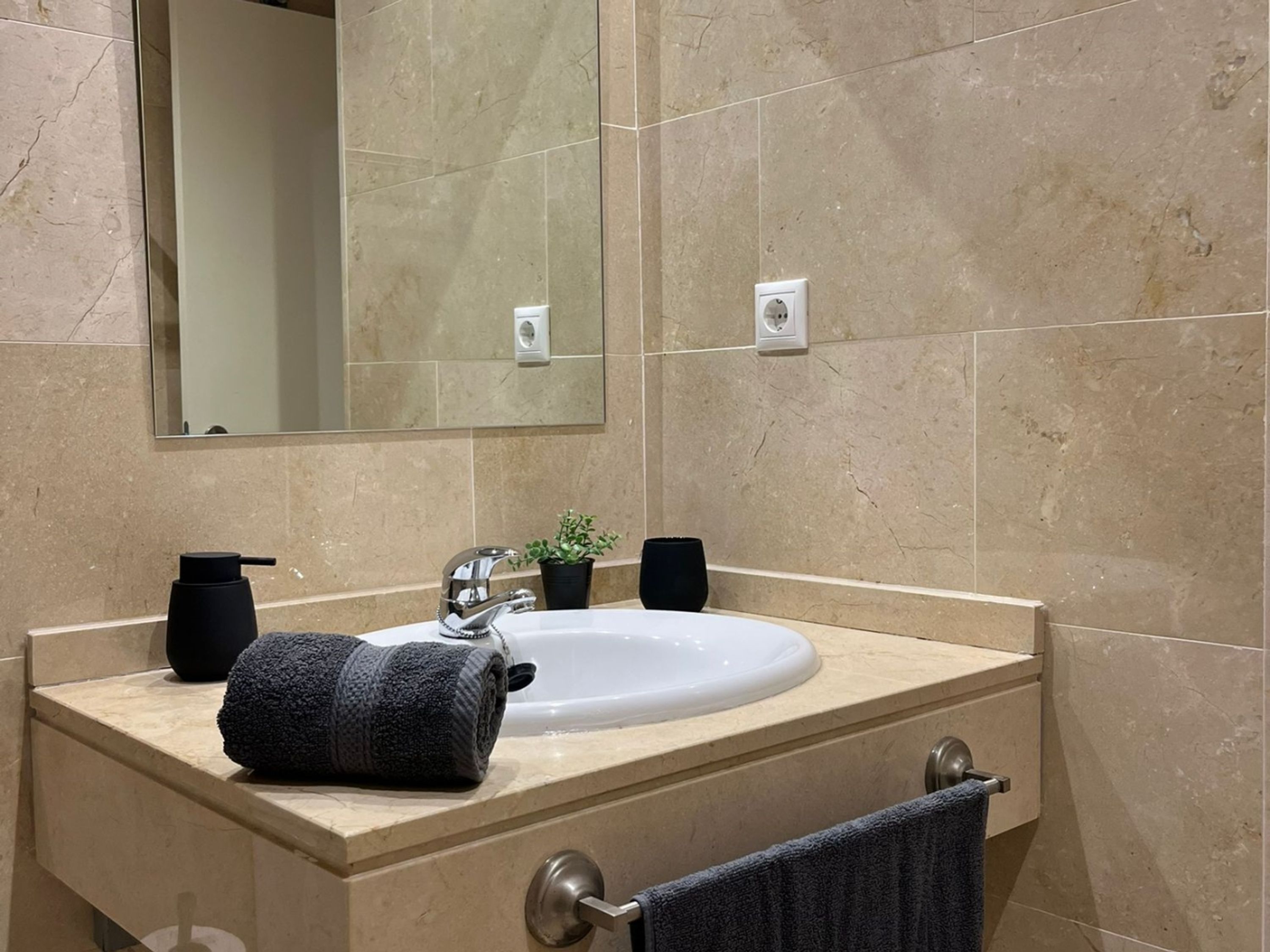 Un baño con lavamanos de mármol color crema, un espejo, una toalla y una pequeña planta en maceta.