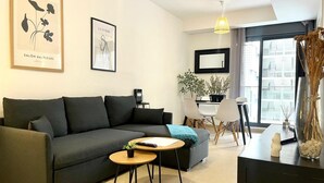 Een moderne woonkamer met een donkergrijze bank, een klein rond houten tafeltje, en een eethoek met een witte tafel en stoelen. Er hangen kunstwerken in lijsten aan de muur, een potplant en een groot raam met uitzicht op gebouwen.