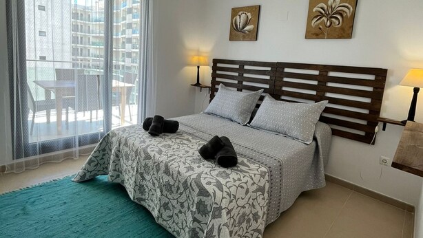 Hotelkamer met een bed, twee wandlampen, een raam met dunne gordijnen en twee bloemschilderijen.