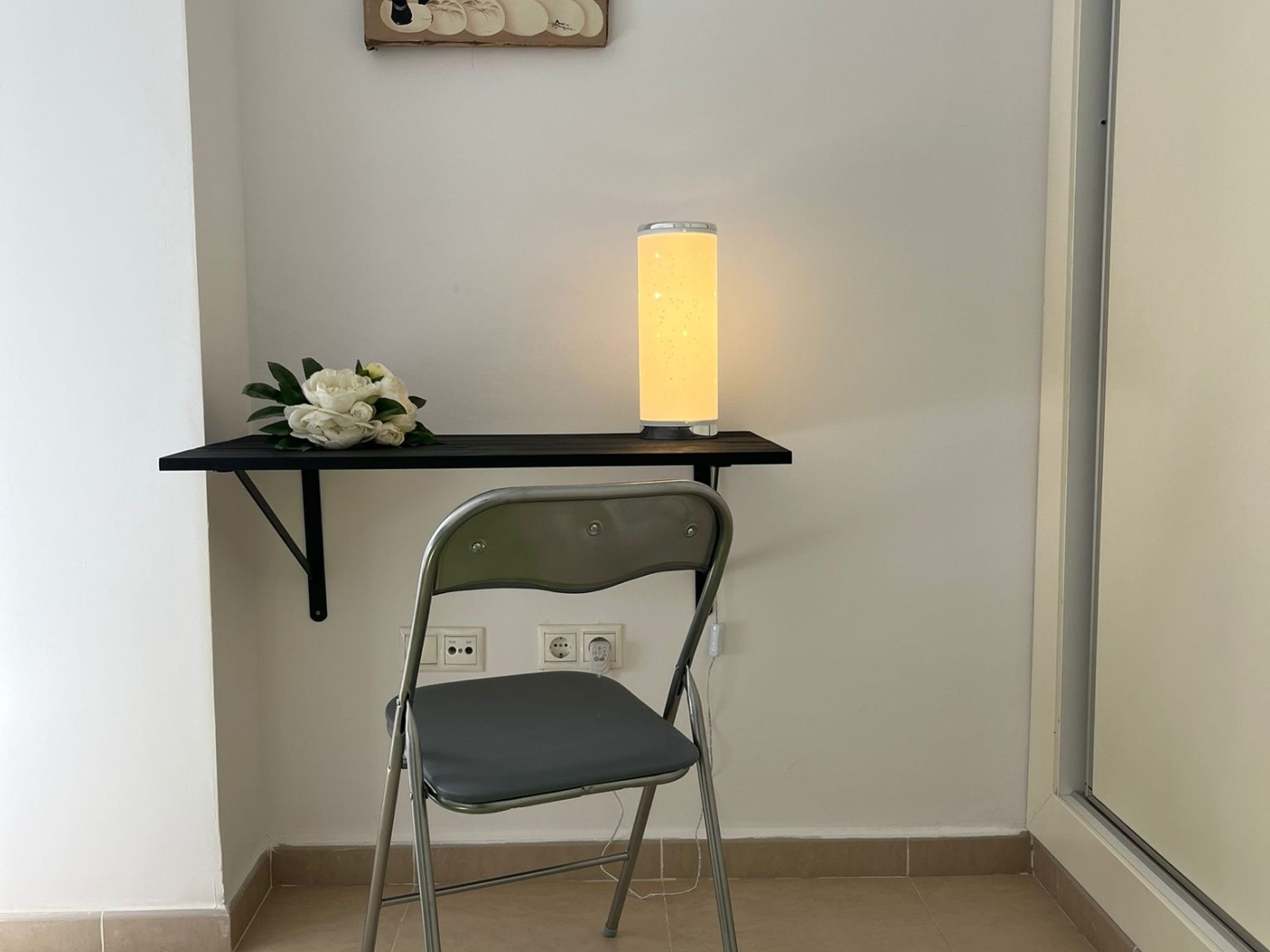 A chair with a metal frame and a dark cushion, a black shelf with a white flower arrangement, and a tall, cylindrical, frosted glass lamp.