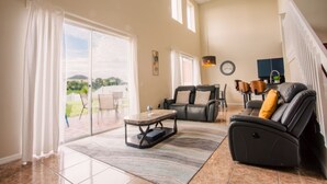 A spacious living room with a sliding glass door leading to a patio, a black leather sofa, a coffee table, and a ceiling light fixture. - Cheerful 5 bed - 4 Bath Spacious Home (Kissimmee)