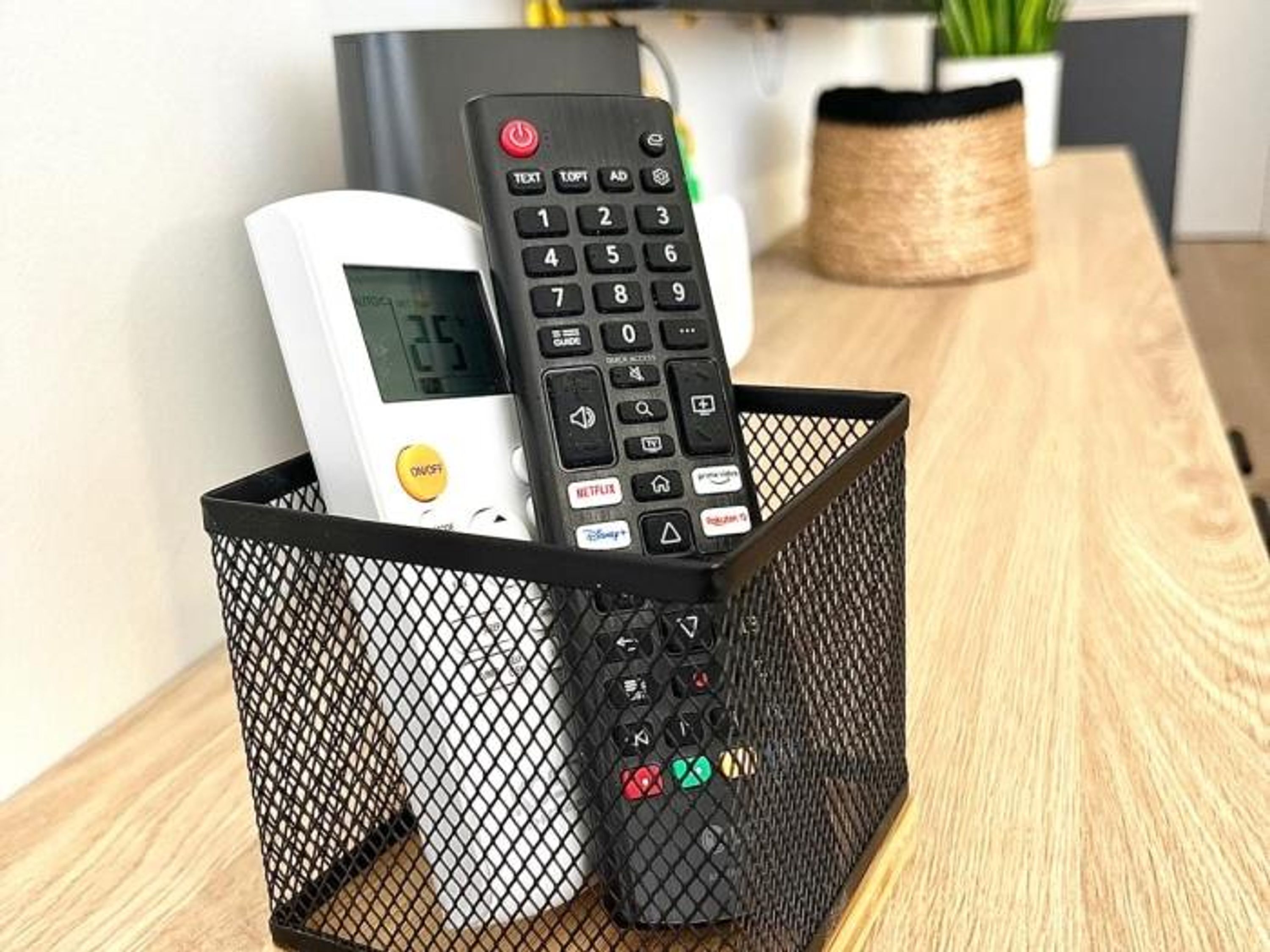 A black mesh basket containing a white digital clock and a black remote control with colorful buttons.