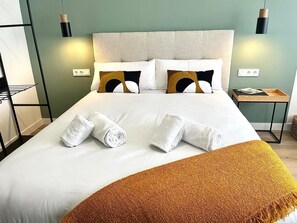 Una cama impecable con sábanas blancas y dos toallas enrolladas, un plaid color mostaza y un cabecero con diseño acolchado.