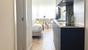Una cocina moderna con encimera negra y electrodomésticos de acero inoxidable. Hay un comedor con una mesa y sillas blancas, y una cama con una colcha amarilla al fondo.