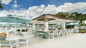 Ein Außenbereich mit Bar, weißen Stühlen, Sonnenschirmen, einem Pool im Hintergrund und Palmen.