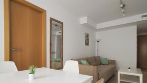 A modern living room with a white dining table, a beige sofa with green cushions, a small white side table, and a wooden door. - La Mar 2 in El Campello (El Campello)