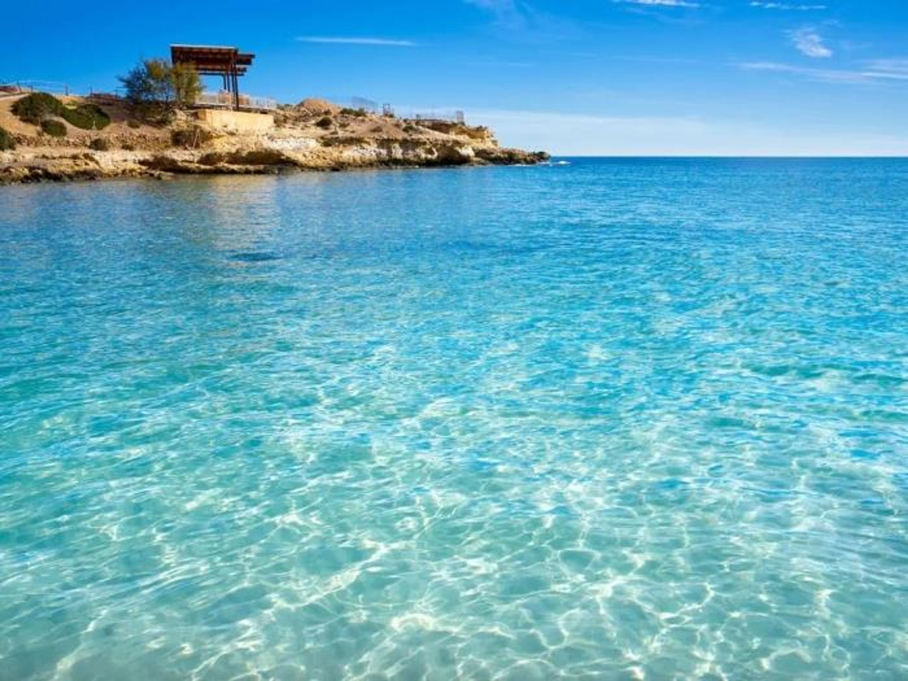 A coastal scene with clear blue water and a rocky outcrop with a wooden structure.