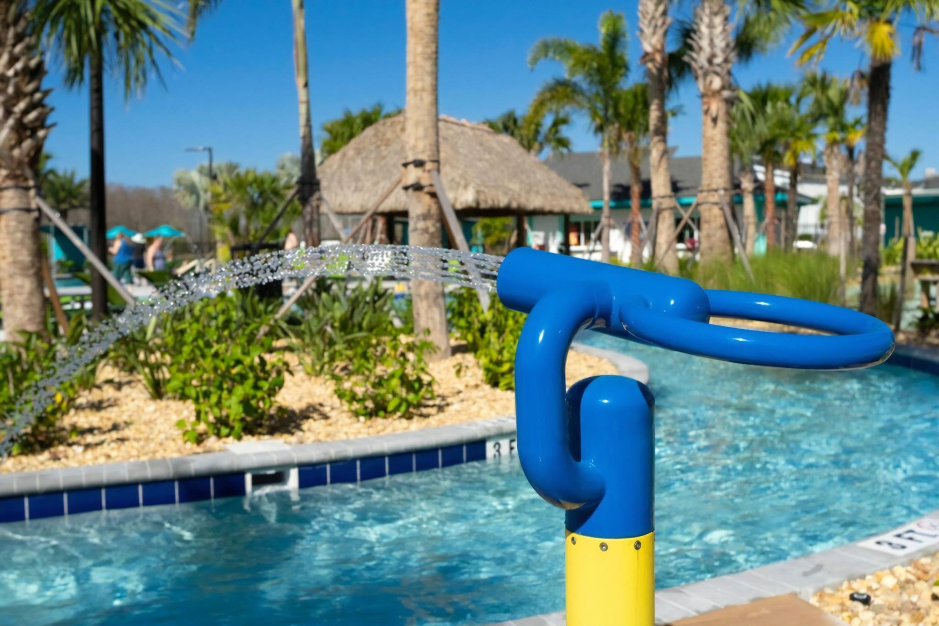 Una piscina con un elemento acuático azul y amarillo, rodeada de palmeras y una estructura con techo de paja.