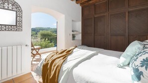 A bedroom with a wooden ceiling, a bed with white bedding, a view of greenery outside, and a balcony with a chair. - Villa Can Fumeral Modern and Seaview Salinas (Sant Antoni de Portmany)