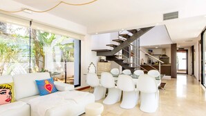 A modern living room with a white sofa, a coffee table, and a unique white pouf. A staircase leads to a kitchen area with a view of the outdoors. - Villa Sa Marinada Modern Villa With Pool for 8 People (Sant Josep de sa Talaia)