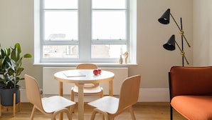 A dining area with a round table, four chairs, a potted plant, and a view of the city through a large window. - Cool Covent Garden 1BR - Netflix Nespresso (London)