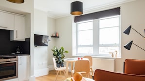 A modern kitchen with white cabinets, a built-in oven, and a black countertop. There is a dining area with a round table, chairs, and a potted plant. A large window provides natural light, and a TV is mounted on the wall. - Cool Covent Garden 1BR - Netflix Nespresso (London)