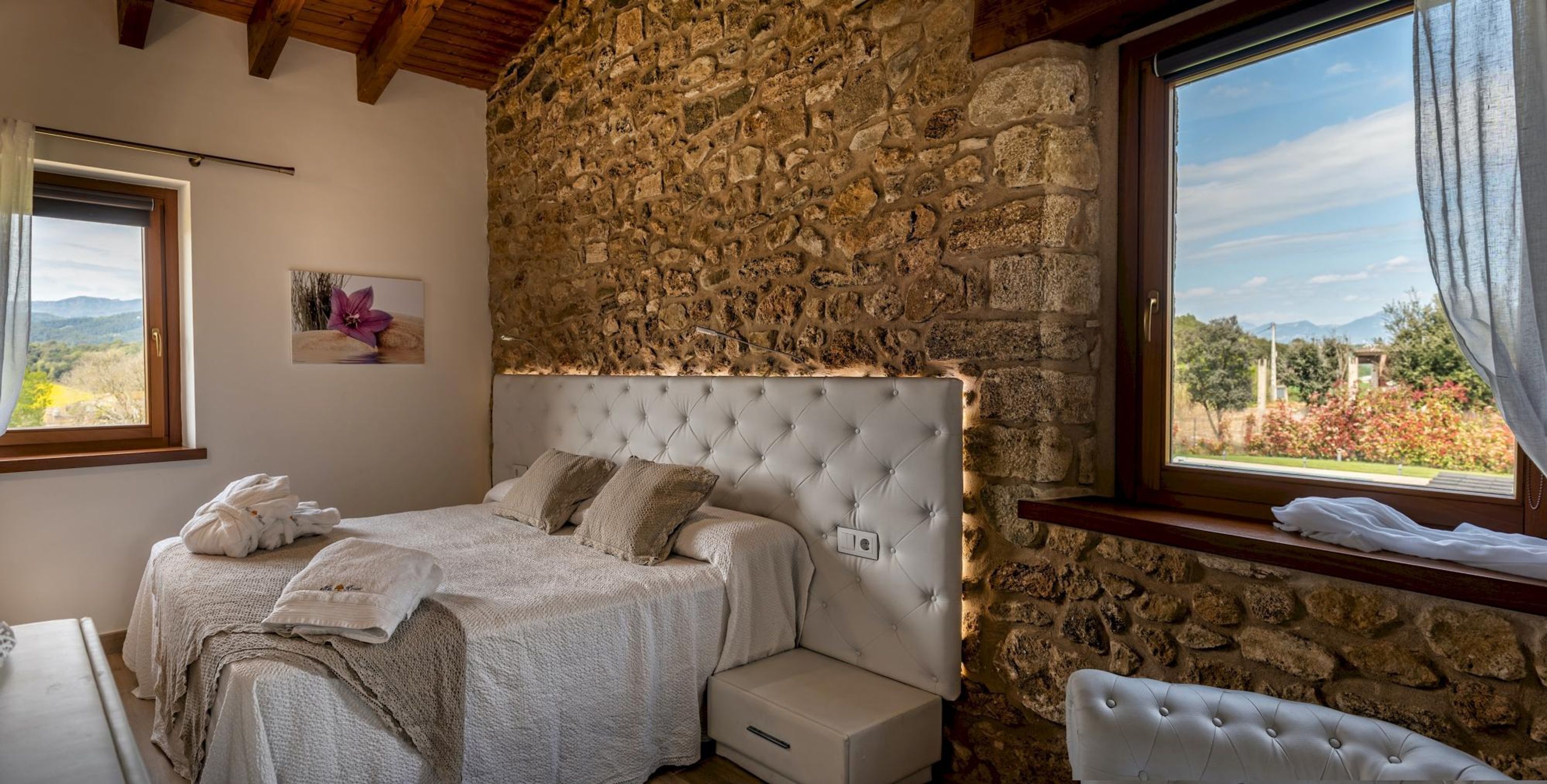 Camera da letto con parete in pietra, una grande finestra e un letto con testiera bianca.