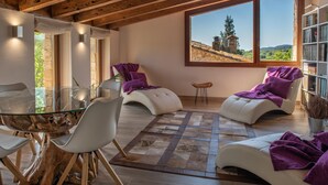 A modern living room with a glass table, white chairs, and a large window with a view of a hill and a tower.