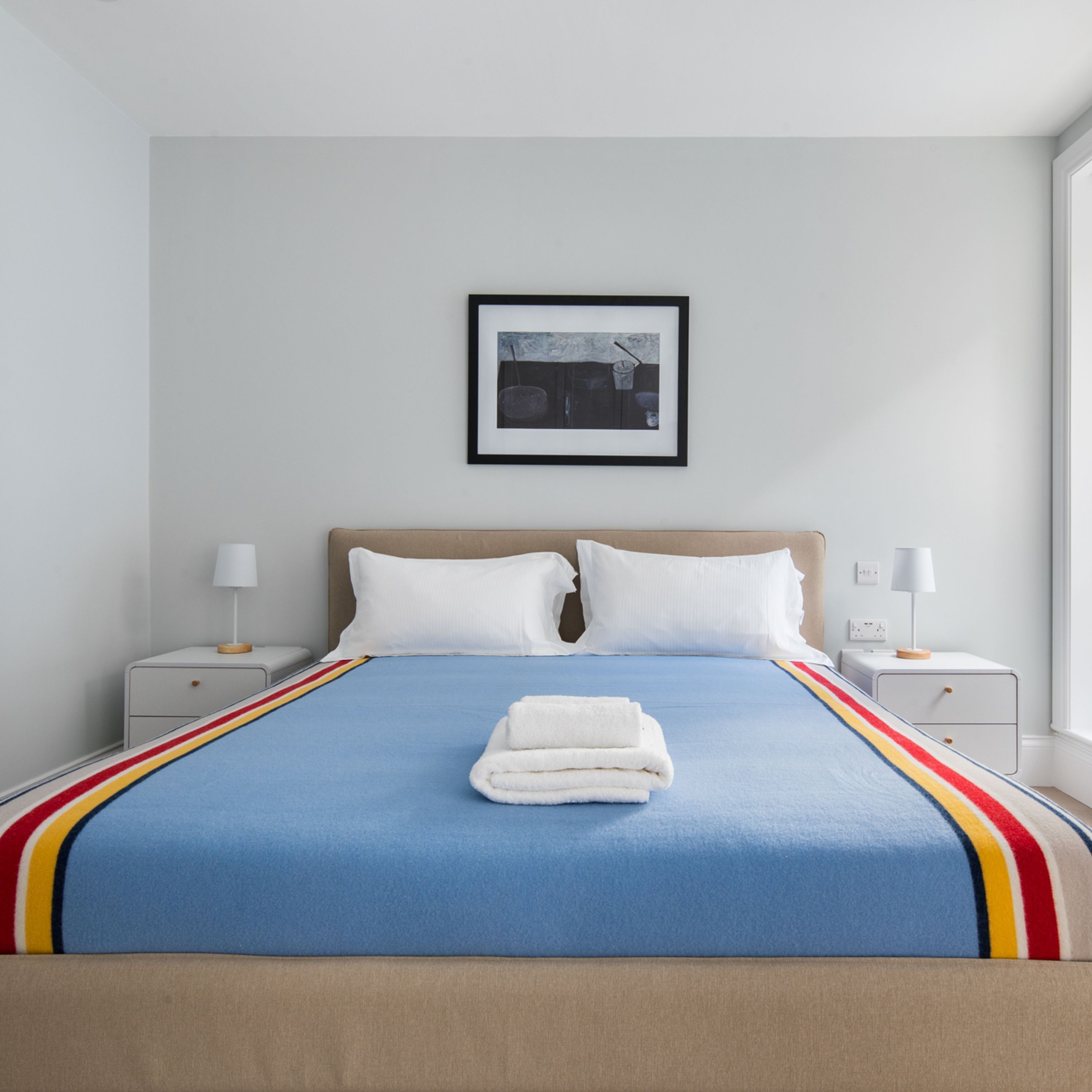 Una cama bien tendida con almohadas blancas y una colcha azul, dos mesitas de noche con lámparas y un cuadro enmarcado en la pared.