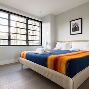 A modern hotel room with a large window, a neatly made bed with white linens, and a framed artwork on the wall.