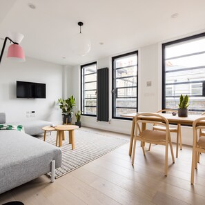 A modern living room with a sofa, a dining table with chairs, a TV mounted on the wall, and large windows with a view of buildings.