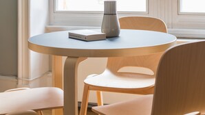 Une table ronde avec un livre et un vase, entourée de deux chaises, avec une fenêtre en arrière-plan.