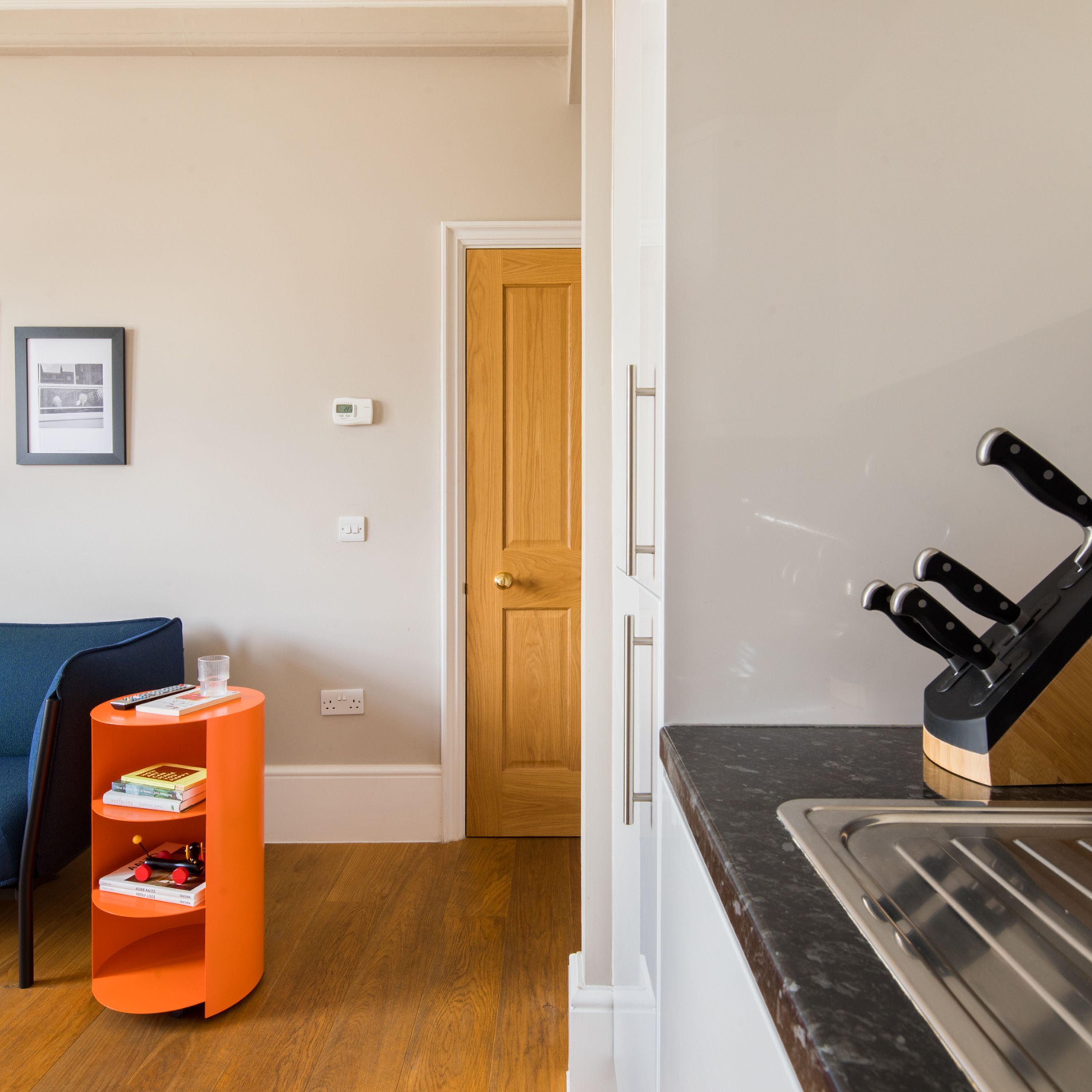 A compact living space with a kitchenette, a wooden door, a framed picture, and a small orange side table with books.