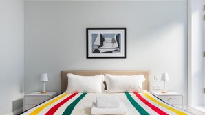 A modern hotel room with a large bed, white linens, a colorful striped blanket, and a framed abstract painting on the wall. - Calm Leicester Sq 1BR - Netflix Nespresso (London)