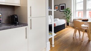 A modern hotel room with a kitchen area, a bed with a black headboard, a dining table with chairs, and a large window providing natural light. - Funky Covent Garden Studio - Netflix Nespresso (London)
