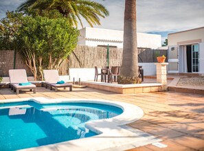 En swimmingpool med solstolar och en palm, omgiven av en vit byggnad med en mur av sten.