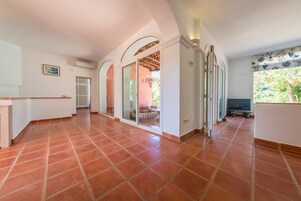 Un pasillo amplio con suelo de baldosas, un portón corredera de vidrio que da a un espacio exterior, y una pequeña zona de estar con una mesa y sillas.