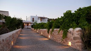 A gravel pathway leading to a white building with a red-tiled roof, surrounded by stone walls and greenery.