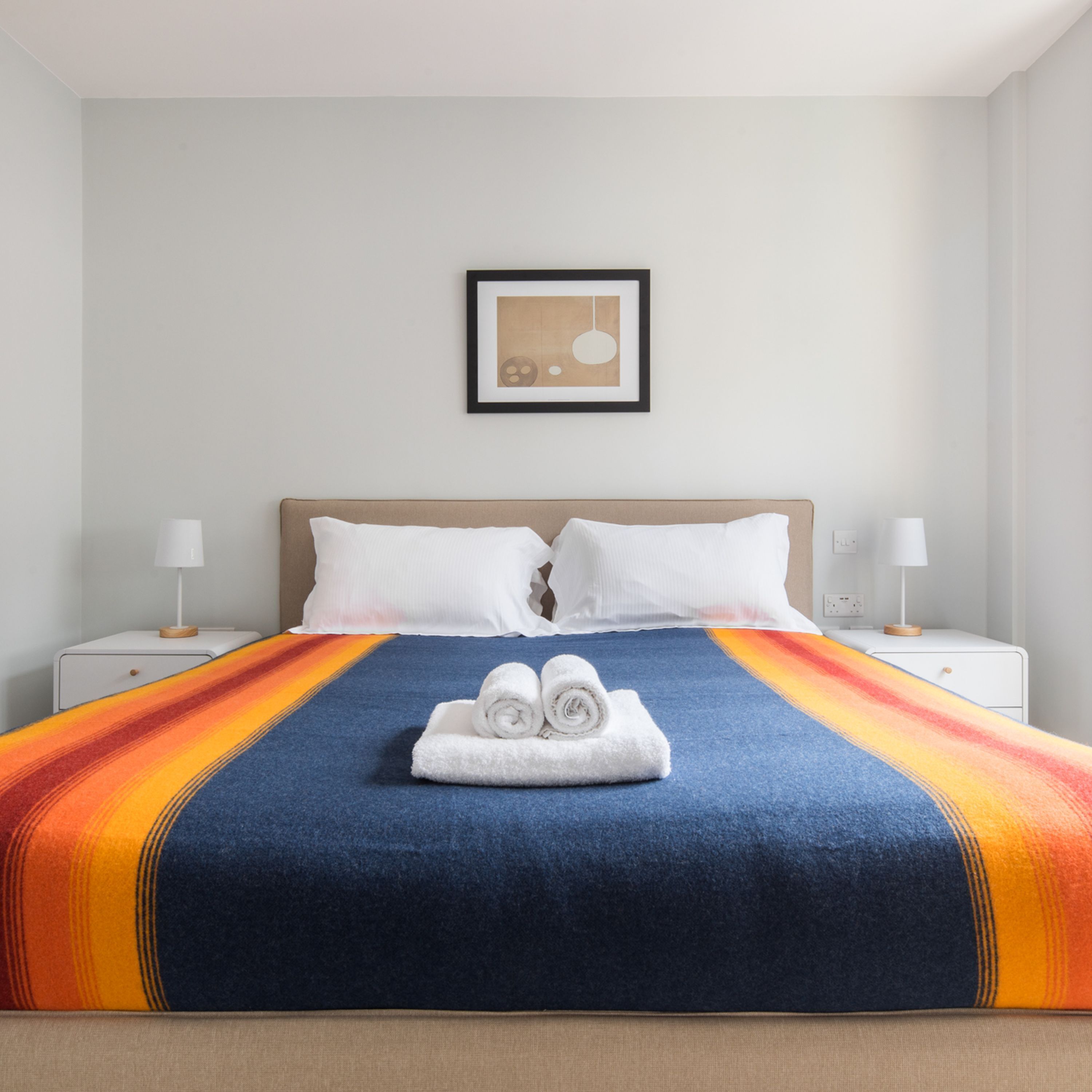 A hotel room with a large bed, two white pillows, a blue and orange striped bedspread, and a framed picture on the wall.