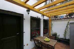 A patio with a wooden table and chairs, a black door, and a window with a metal gate and red flowers.