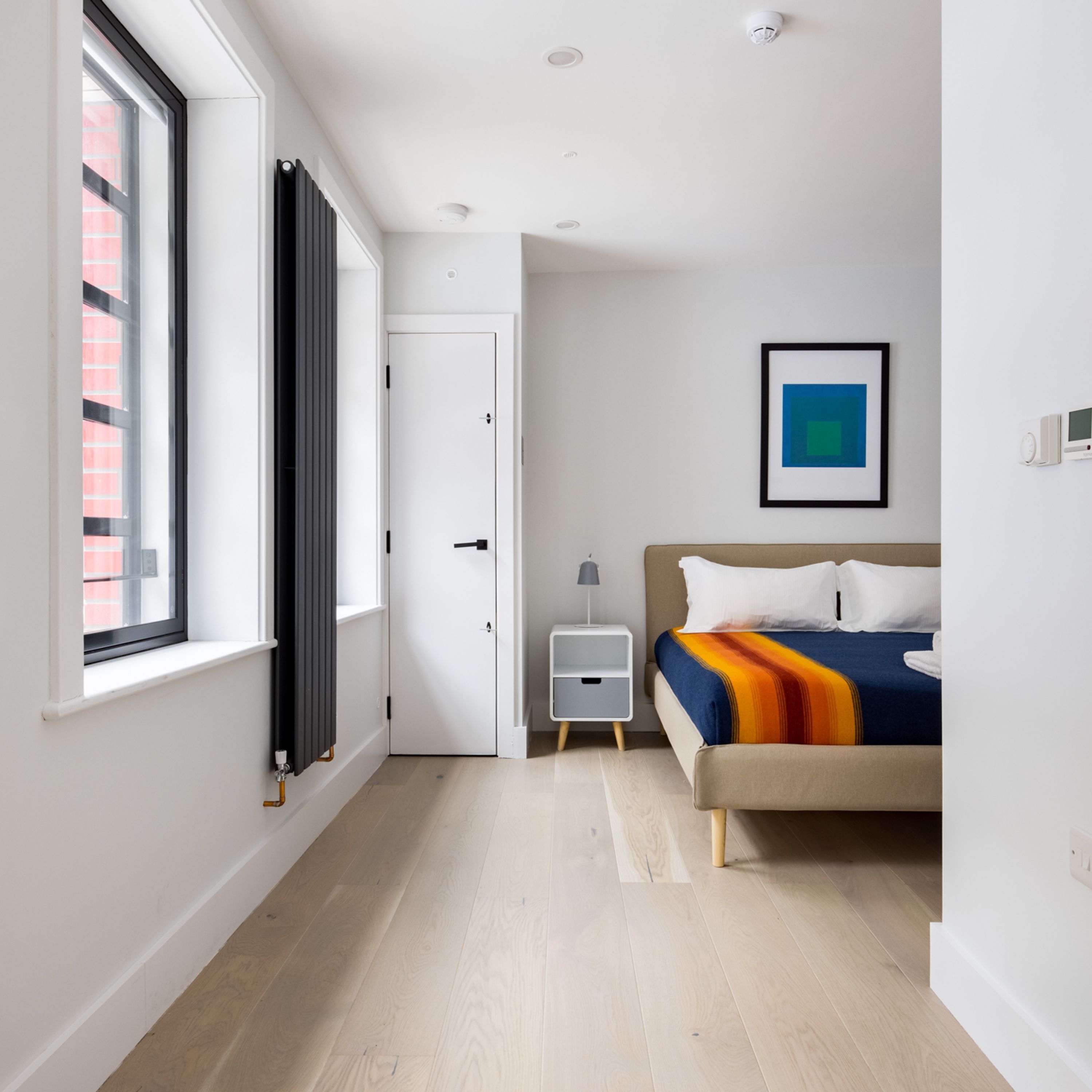 Une chambre à coucher moderne avec une grande fenêtre, un lit recouvert d’une couverture colorée, une table de chevet avec une lampe et un tableau encadré accroché au mur.