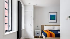 A modern bedroom with a large window, a bed with a colorful blanket, a bedside table with a lamp, and a framed picture on the wall. - Cool Leicester Sq Studio - Netflix Nespresso (London)