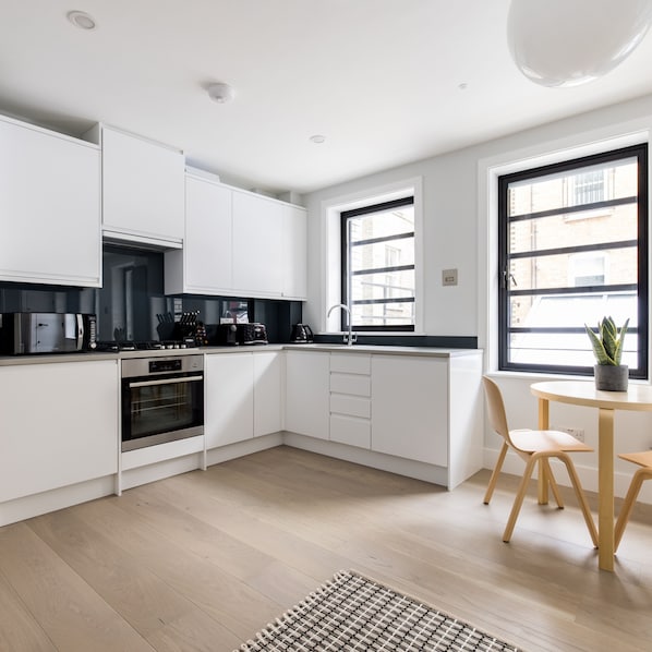 A modern kitchen with white cabinets, a built-in oven, a coffee machine, and a dining area with a table and chairs.