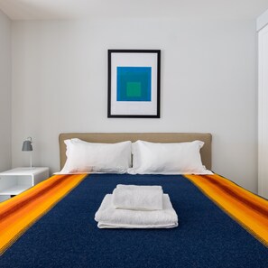 A hotel room with a bed, white pillows, a blue and orange striped bedspread, a folded white towel, a bedside lamp, and a framed picture on the wall.