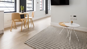 A modern, well-lit room with a flat-screen TV mounted on the wall, a dining area with a table and chairs, and large windows offering a view of the city. - Cool Leicester Sq Studio - Netflix Nespresso (London)