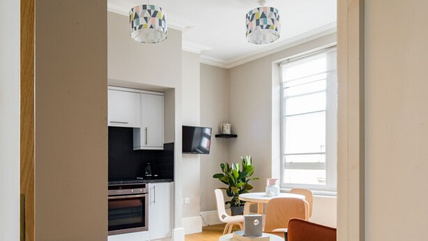 A modern kitchen with white cabinets, a black countertop, and a built-in oven. There is a dining area with a table and chairs, a potted plant, and a small white table. Two pendant lights hang from the ceiling.