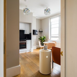 Een moderne keuken met witte kasten, een zwart aanrecht en een inbouwoven. Er is een eethoek met een tafel en stoelen, een potplant en een kleine witte tafel. Twee hanglampen hangen aan het plafond.