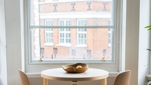 Une pièce avec une table ronde en bois et deux chaises, une fenêtre donnant sur un bâtiment en briques, et une plante sur la droite.