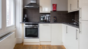 Une cuisine moderne avec des meubles blancs, une hotte de cuisinière en acier inoxydable, un four encastré, une machine à café et une couverture anti-feu accrochée au mur.