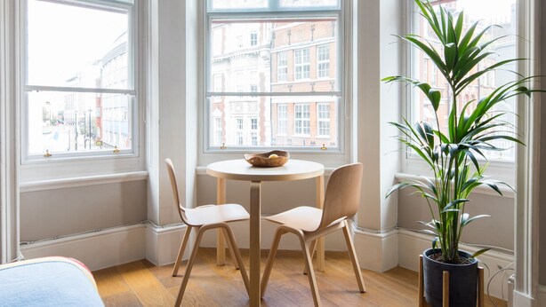 Une pièce d’angle avec une baie vitrée, une table ronde en bois avec deux chaises, une plante en pot et un lit au premier plan.