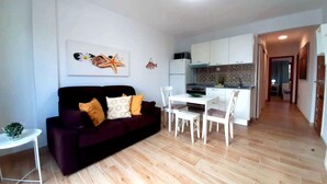 Un espai vital compacte amb un sofà de color fosc, una taula de dinar petita amb cadires, una zona de cuina amb armaris blancs i un respoldre de rajola.