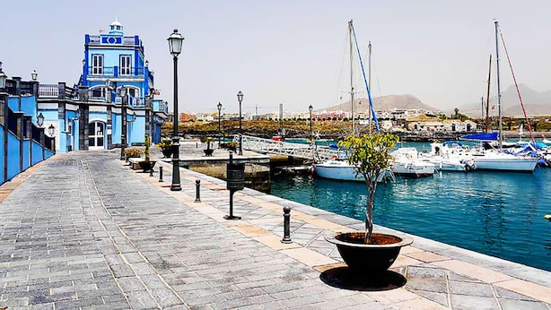 A waterfront promenade with a blue building, potted plants, and boats docked in the background. - Las Galletas Ocean Plata - 2D (Arona)