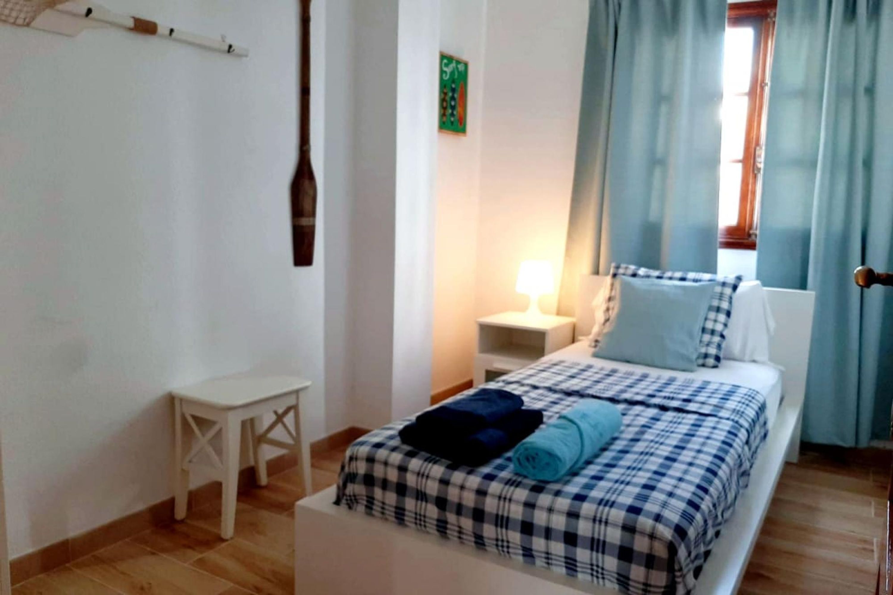A bedroom with a bed, a small table, a nightstand, a lamp, and a window with curtains.