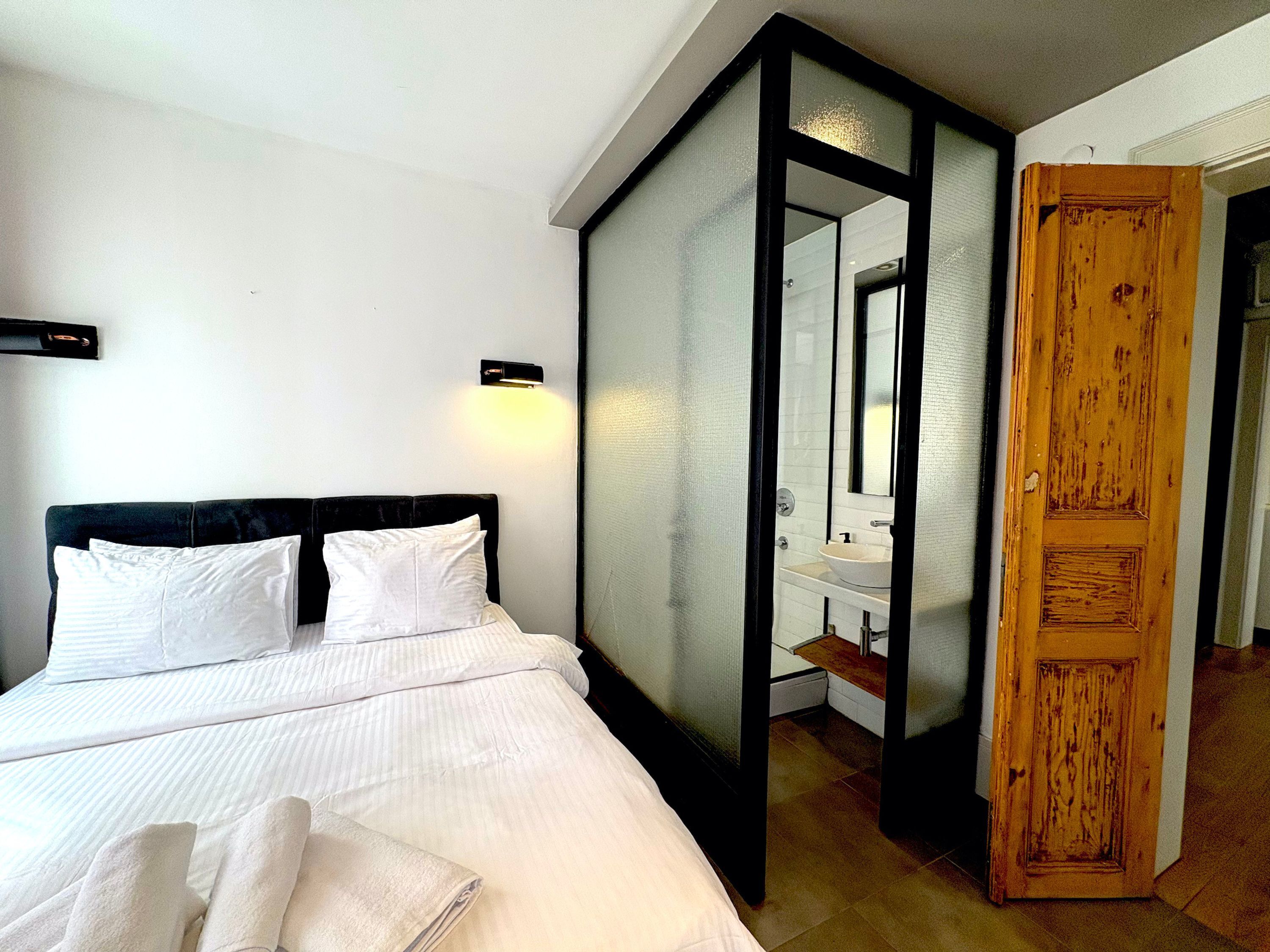 A bedroom with a bed, white linens, a nightstand, a wall-mounted light, and a wooden door leading to a bathroom with a sink and mirror.